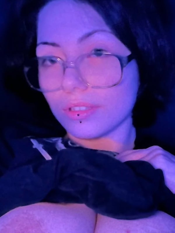 Young woman with dark hair and glasses smiling at the camera under purple lighting, wearing a dark top.