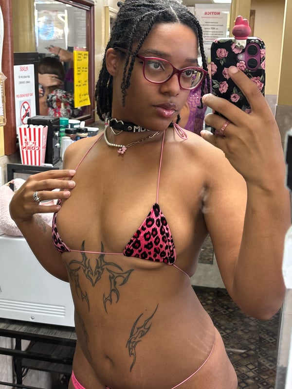 Young woman with braided hair and glasses taking a mirror selfie in a bathroom, wearing a colorful animal print top and necklace.
