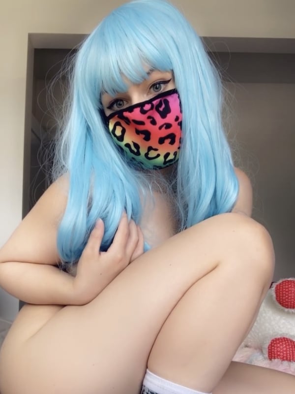 A young woman with blue hair and a leopard print face mask sits with knees up in a room with plush toys nearby, wearing white socks.