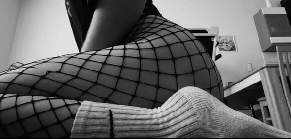 Close-up view of a person sitting on a bed wearing fishnet stockings and a white sock, in a cozy bedroom setting with furniture and decor.