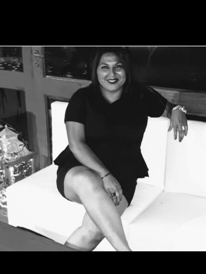 A smiling woman with dark hair sitting on a white couch, dressed in a black top and skirt, with a window and decor in the background.
