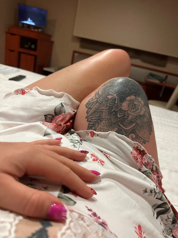 Close-up of a person's leg with intricate floral tattoos, resting on a bed in a cozy bedroom with furniture and a TV in the background.