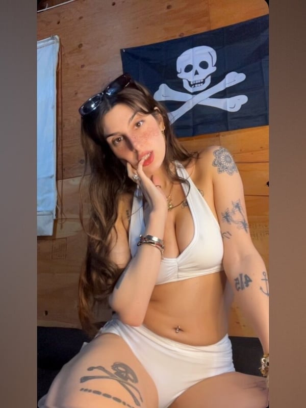 Young woman with sunglasses on her head, wearing a white halter top and shorts, posing in front of a pirate flag against a wooden wall.