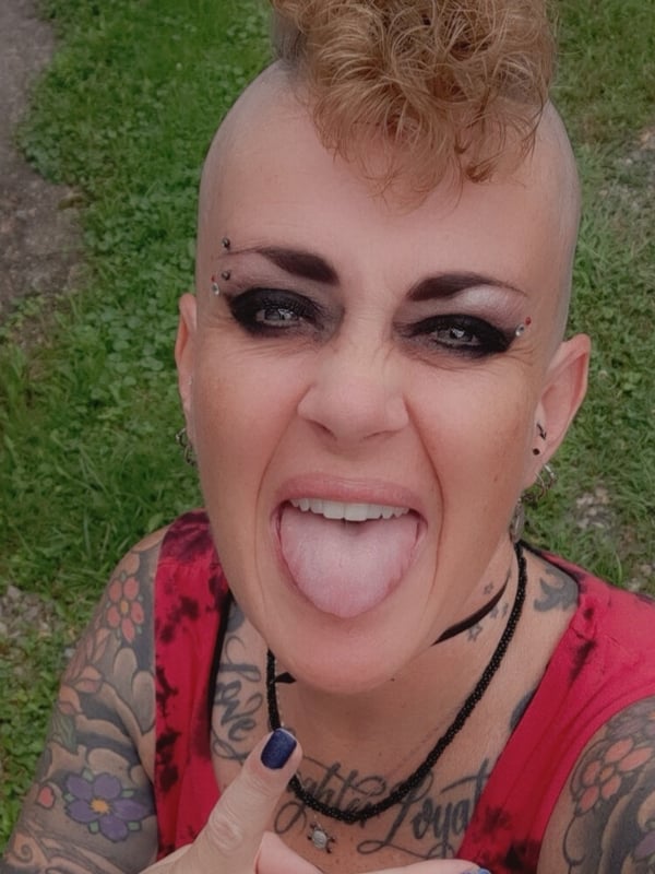 A woman with a distinctive mohawk and facial piercings stands outdoors on grass, wearing a red top, sticking out her tongue playfully.