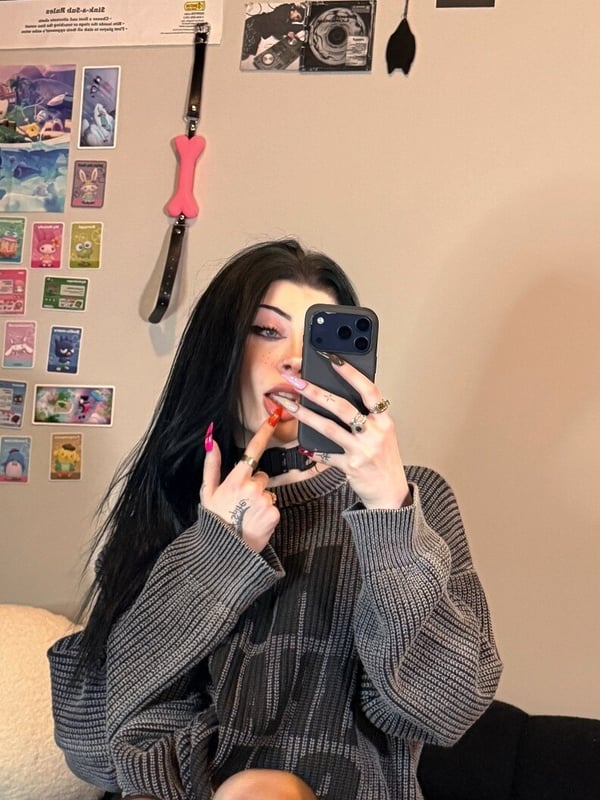 A person with long dark hair and glasses taking a mirror selfie in a cozy room with anime posters and decorations on the wall.