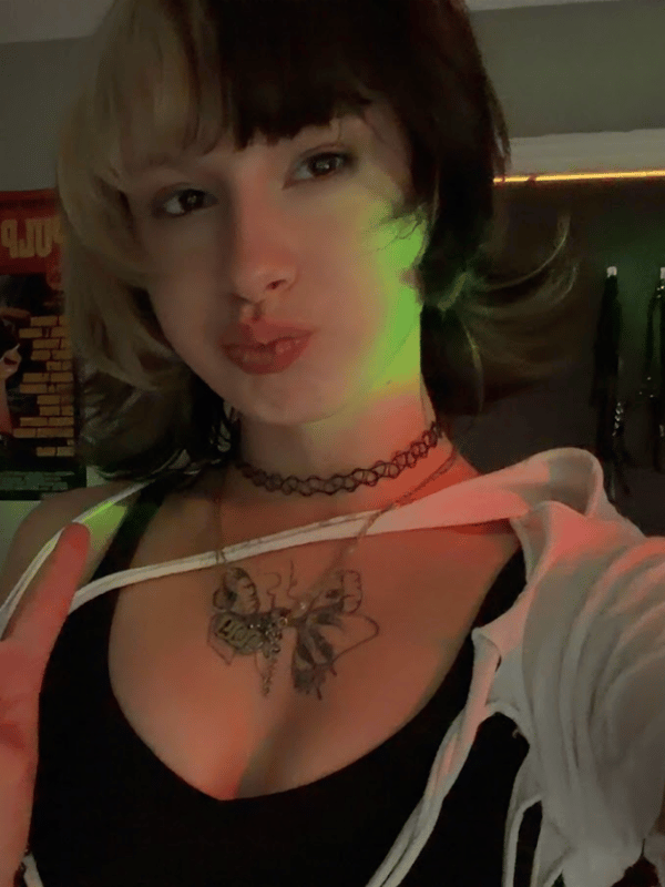 A young woman with split black and blonde hair making a peace sign, wearing a choker and open hoodie with tattoos on her chest, standing indoors with green lighting.