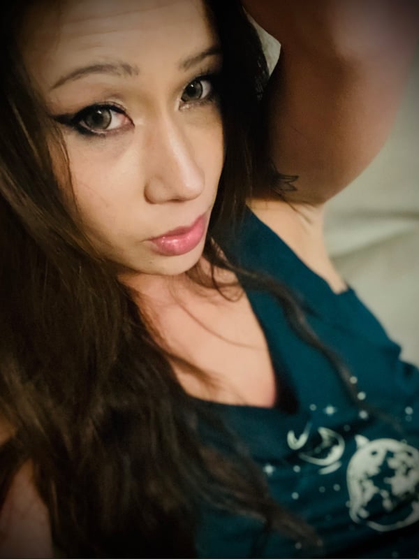 Young woman with long brown hair and striking makeup, close-up portrait wearing a sleeveless top, gazing at the camera.