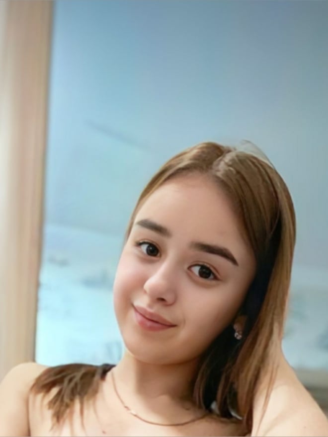 Young woman with long brown hair smiling softly at the camera, standing in front of a window with light wooden frames.
