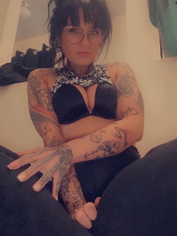 A woman with dark hair in a bun poses in a bathroom setting, wearing glasses, a black choker, and a black top with intricate tattoos on her arms.