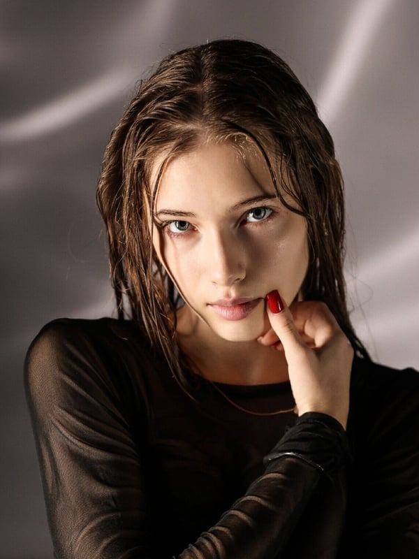 Young woman with wet brunette hair wearing a sheer black top, posing thoughtfully with a finger to her lips against a soft gray backdrop.