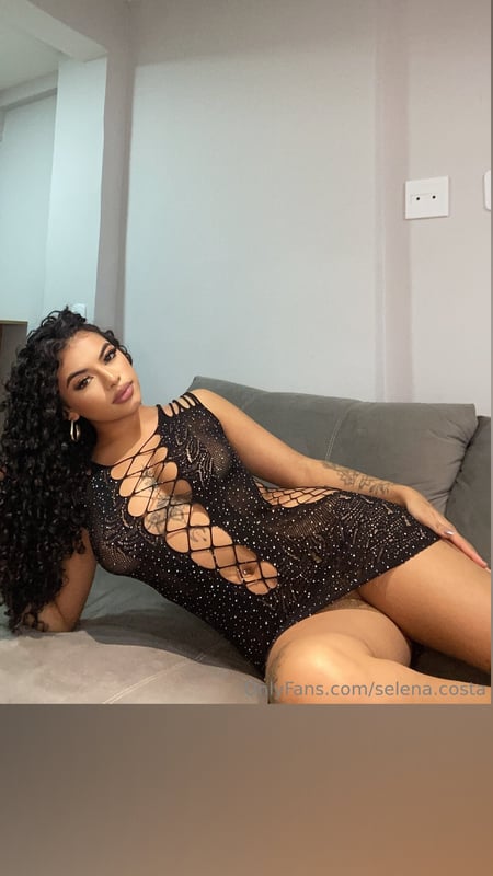 A woman with curly hair lounging on a couch in a stylish black lace outfit against a neutral background.
