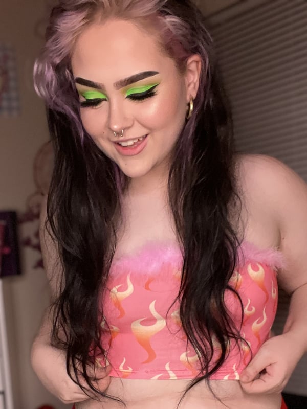 Young woman with long dyed pink and black hair, green eyeshadow, and a smile, posing in a fuzzy orange and pink tube top in a cozy bedroom setting.