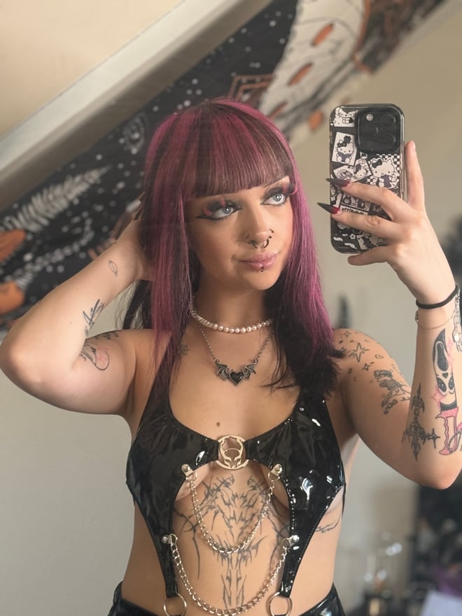 A tattooed person with dark magenta hair and bangs posing indoors in patterned clothing, with colorful ambient lighting in the background.
