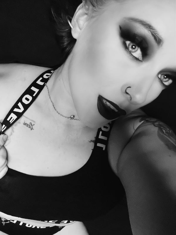 Close-up portrait of a woman with blonde hair, heavy makeup, and piercings, wearing a black top and necklace against a dark background.