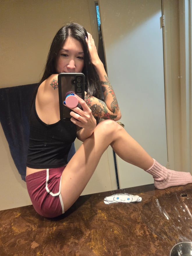 A woman with long dark hair taking a mirror selfie in a bathroom, wearing a black tank top and maroon shorts while sitting on the floor.