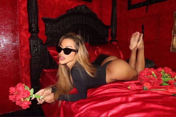 A woman posing on a red satin bed in a black outfit with sunglasses, holding red roses in a luxurious bedroom setting.