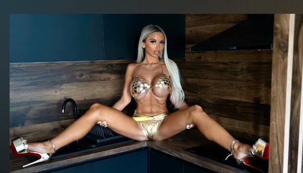 A woman posing in a kitchen area with wooden walls and modern decor, wearing shiny metallic attire and high heels.