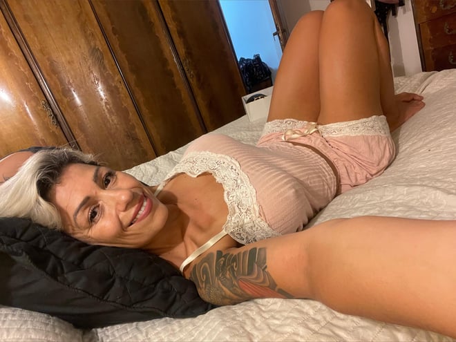 A woman with platinum blonde hair reclining on a bed in pink lace pajamas, smiling at the camera in a cozy bedroom setting.