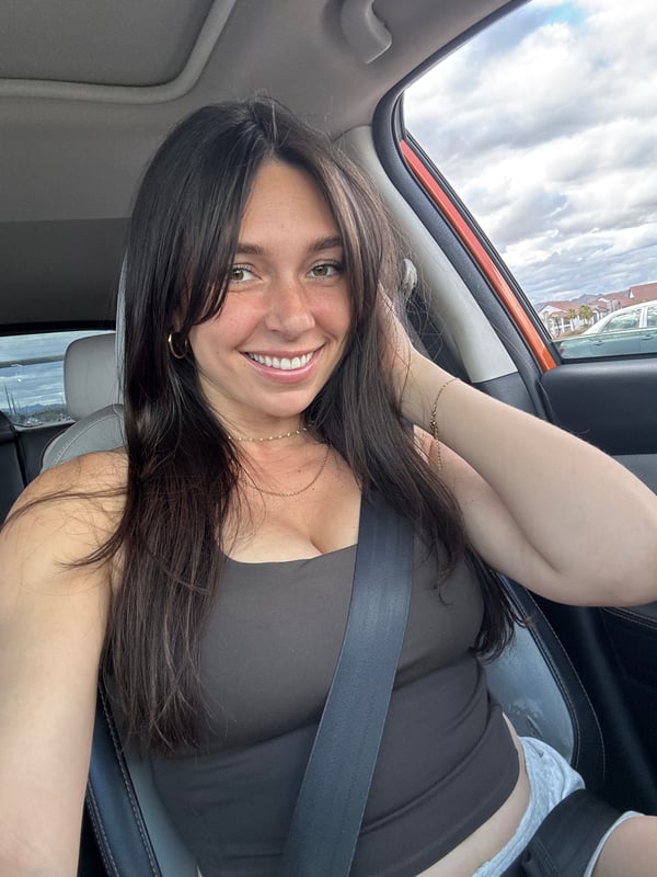 A woman with long dark hair sitting in a car, smiling softly at the camera with a scenic outdoor background visible through the window.