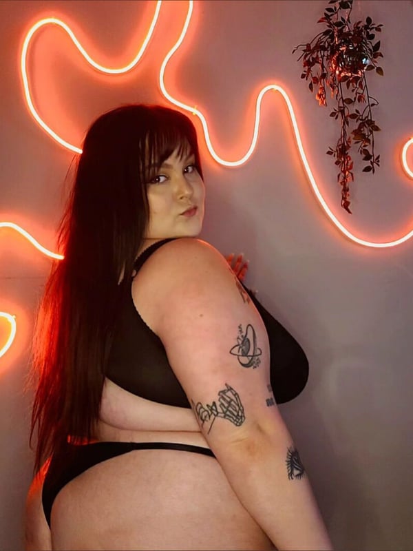 A woman with long dark hair and tattoos posing sideways in black lingerie against a neon-lit wall with plants.