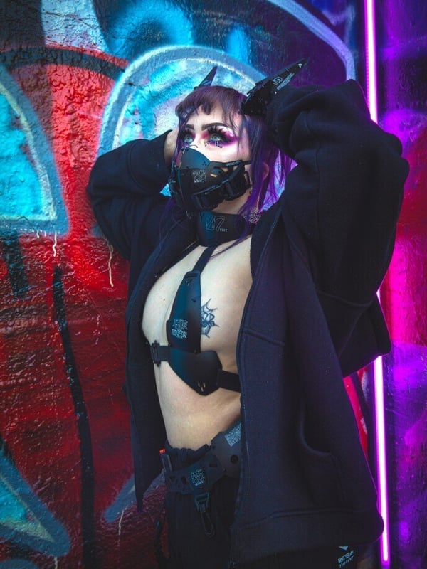 Woman with purple hair wearing a harness and mask, posing against a graffiti wall with neon lights in an urban cyberpunk style.