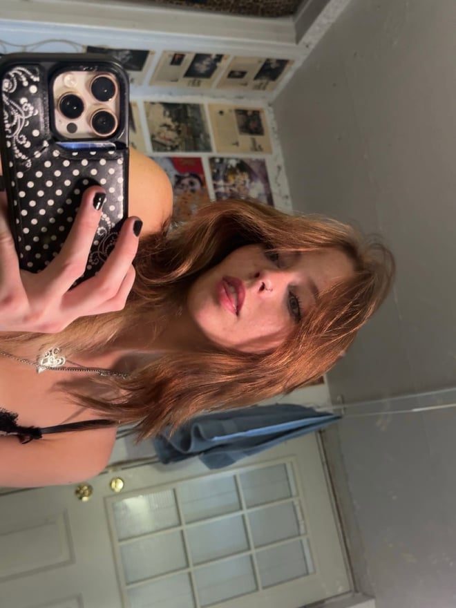 Close-up portrait of a young woman with brown hair and facial piercings, posed in a bedroom with colorful artwork.