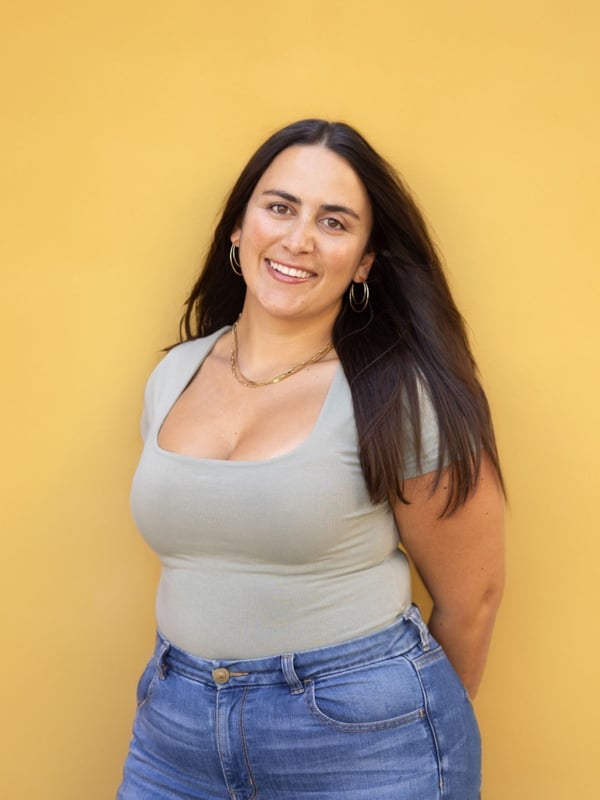 A smiling woman with long dark hair posing confidently in a gray tank top and blue jeans against a solid yellow background.