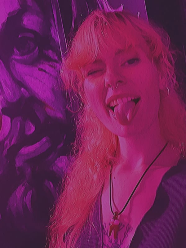 A young woman with long wavy red hair sticking out her tongue, smiling playfully against a vibrant purple artistic background.