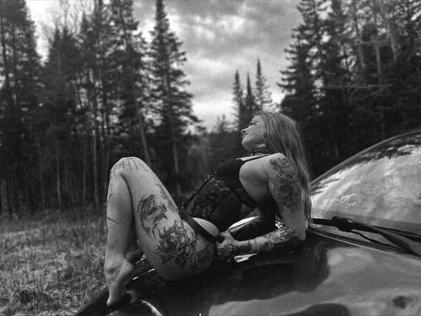 A woman with long hair and tattoos reclines on a car hood in a forested area under a cloudy sky, wearing dark lingerie.