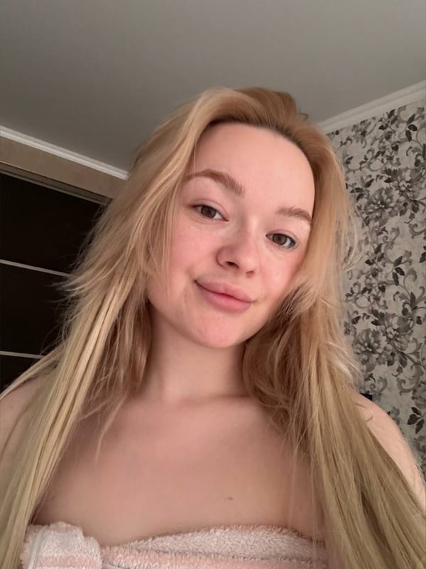 Close-up selfie of a young woman with long blonde hair smiling softly, wrapped in a towel inside a room with floral wallpaper.