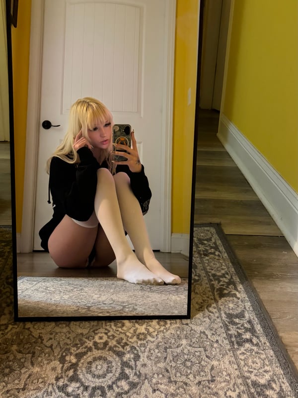 Young woman with long blonde hair taking a mirror selfie in a bikini top, holding a phone, with tattoos on her arm and a beach-themed background.