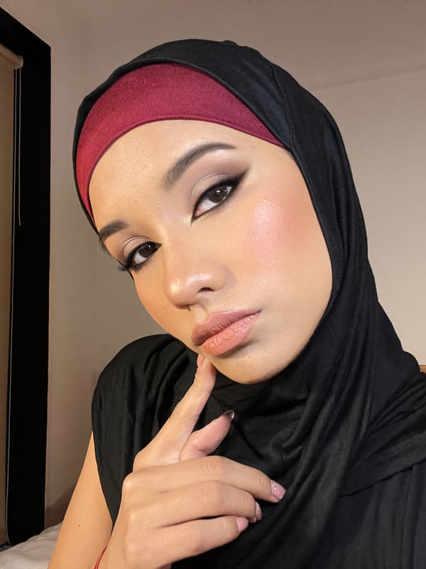 A close-up selfie of a woman wearing a hijab, posing indoors with her hand near her face.