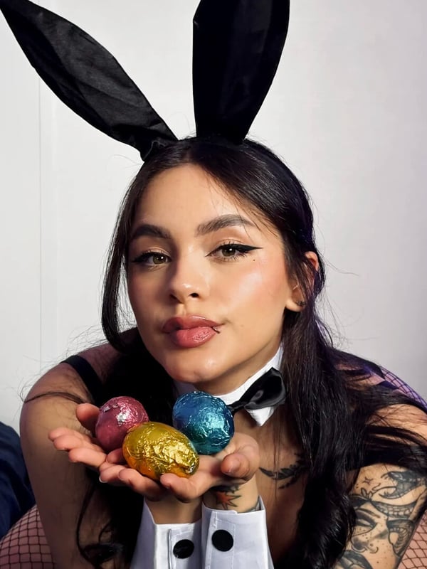A woman with long dark hair, tattoos, and bunny ears holding colorful eggs while wearing a black bow tie and fishnet outfit.