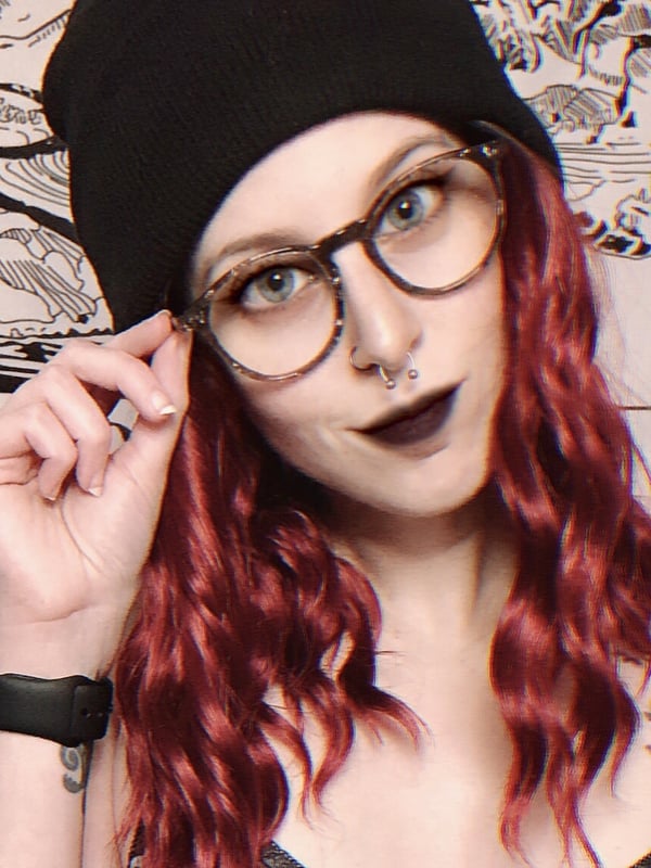 A woman with red curly hair, black beanie, glasses, and dark lipstick, posing playfully indoors with a tapestry background.