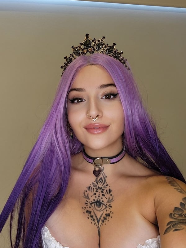 A woman with long purple hair wearing a crown and choker necklace, posing in a white corset top indoors.