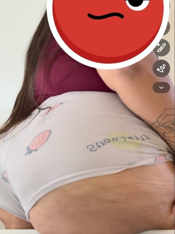 Close-up of a woman's backside in white underwear with tattoos visible, overlaid on a red Snapchat filter with a fierce expression emoji, shown on a mobile screen.
