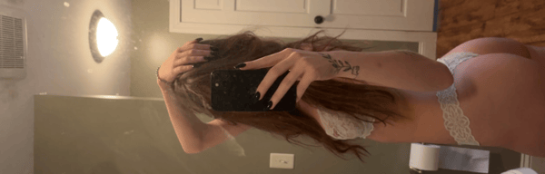A person taking a mirror selfie in white lace underwear, showing back and side profile with long brown hair.