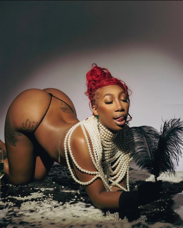 A woman with red hair in a bun poses on the floor wearing a pearl necklace, black gloves, and a feather accessory against a dark background.