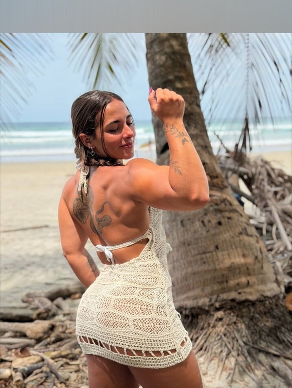 Athletic woman flexing her muscles on a sunny beach next to a palm tree, wearing a crocheted dress.
