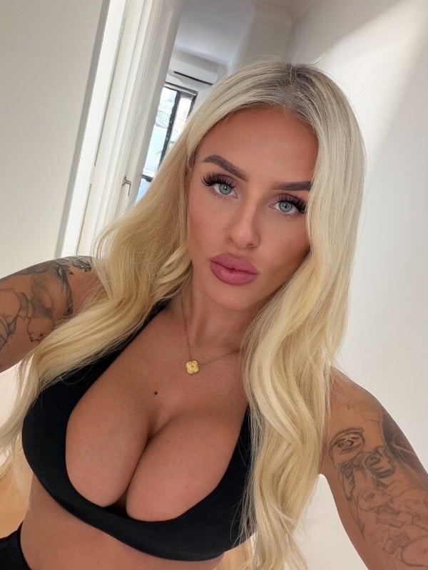 A woman with long blonde hair taking a selfie in a bright hallway, wearing a black crop top and gold necklace.