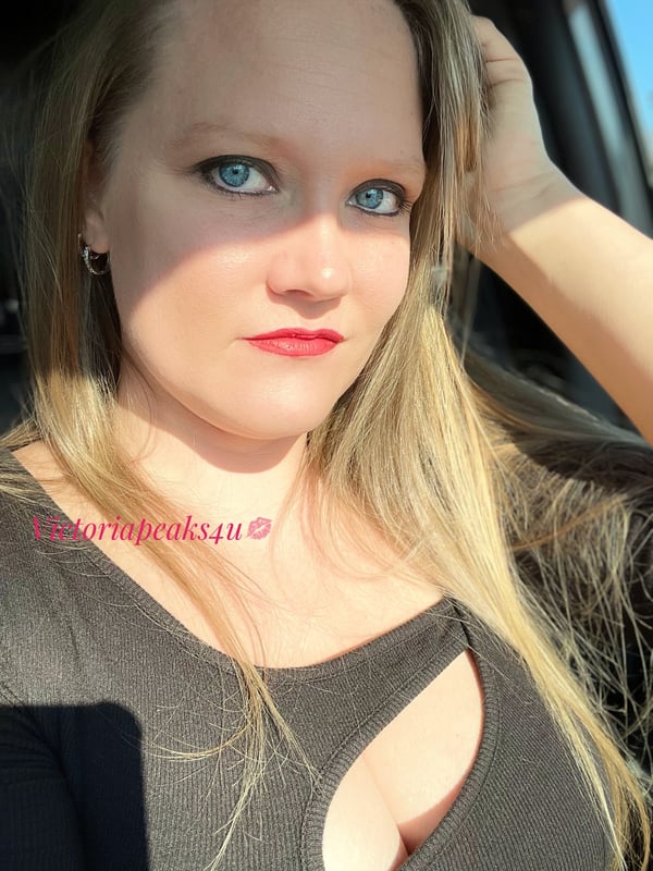 A woman with long blonde hair and blue eyes poses confidently in a car, wearing a black top with cutouts.