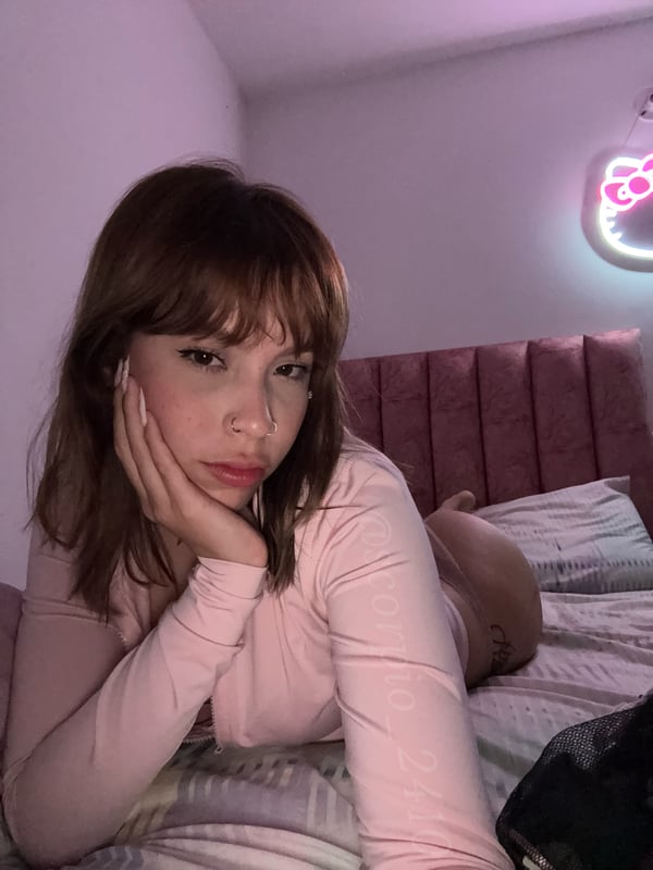 A young woman with a thoughtful expression resting her face on her hand in a pink bedroom with neon lighting.