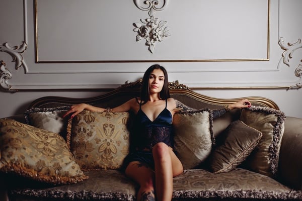 A young woman with long dark hair sits on an ornate vintage sofa in a luxurious room with decorative walls, wearing a black lace top.