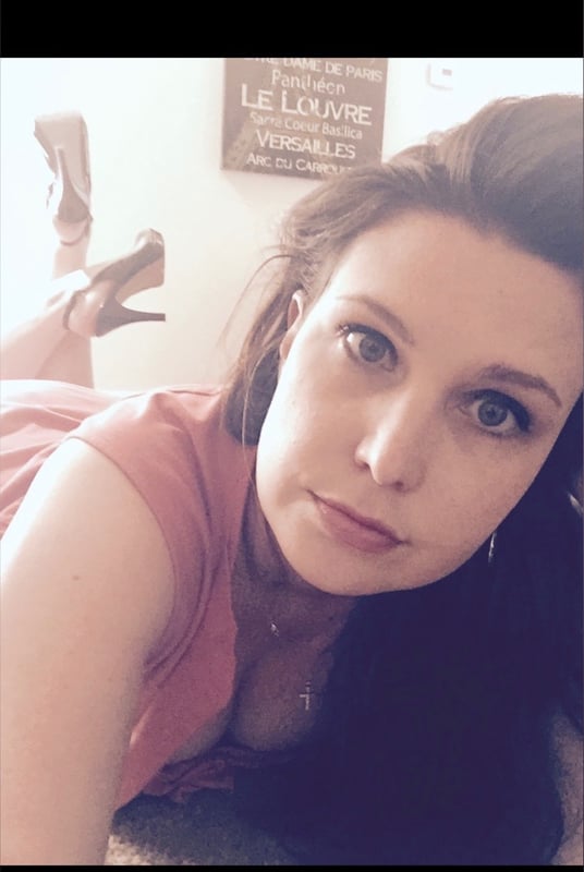 Woman with long brunette hair reclining on a bed in a pink sleeveless top and high heels, with a decorative sign on the wall behind her.