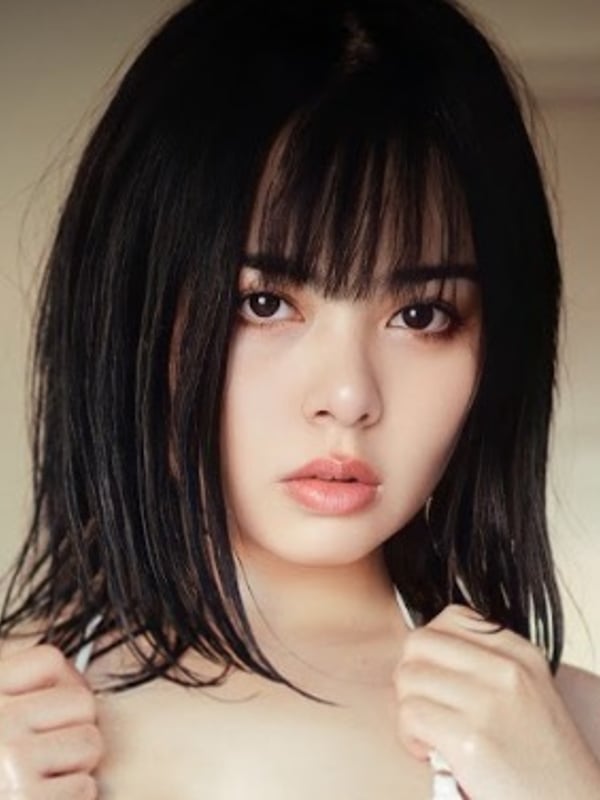 Young woman with straight black bangs smiling softly at the camera, wearing a white top.