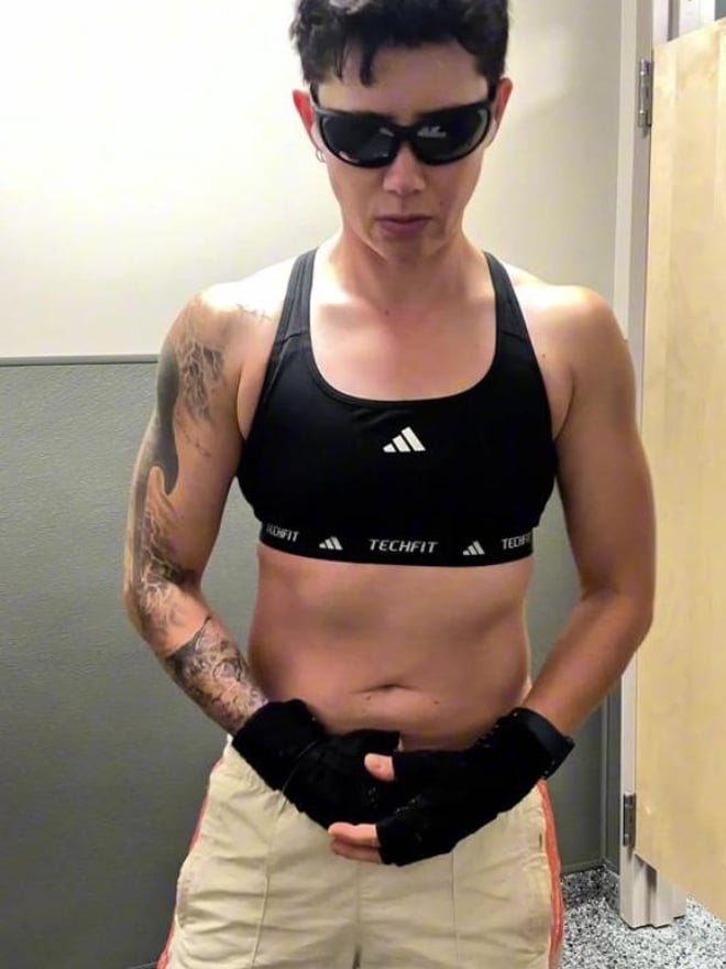 A muscular person wearing a cowboy hat and harness flexes in front of a dark textured wall.