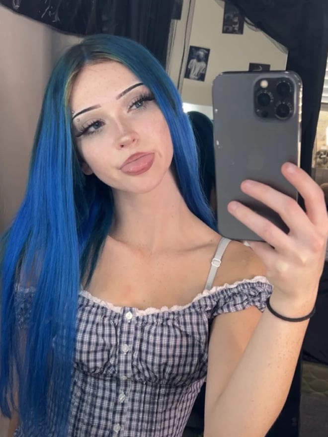 A young woman with long blue hair posing for a mirror selfie in a checked top.