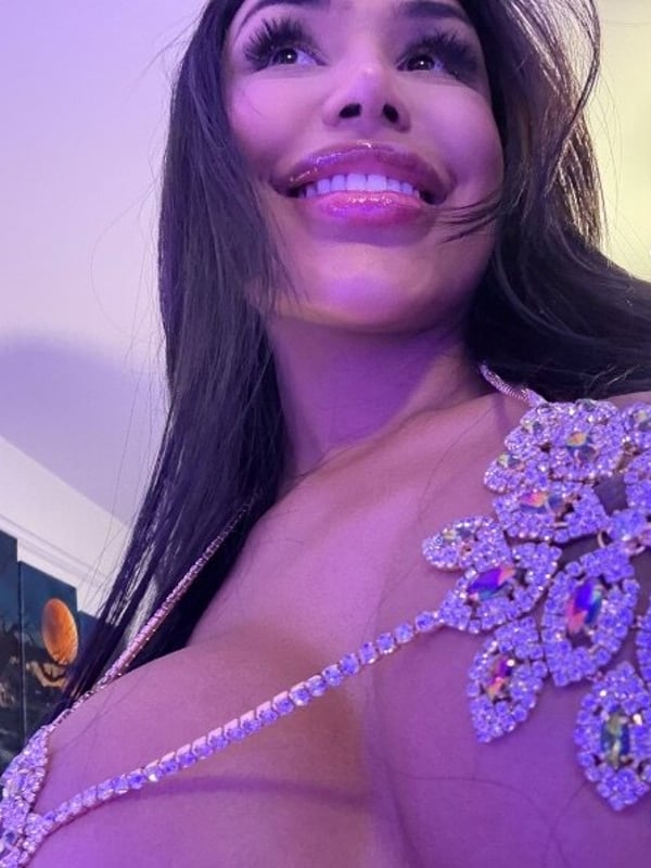 A smiling woman with long dark hair wearing a glittering top, illuminated by purple lighting in an indoor setting.