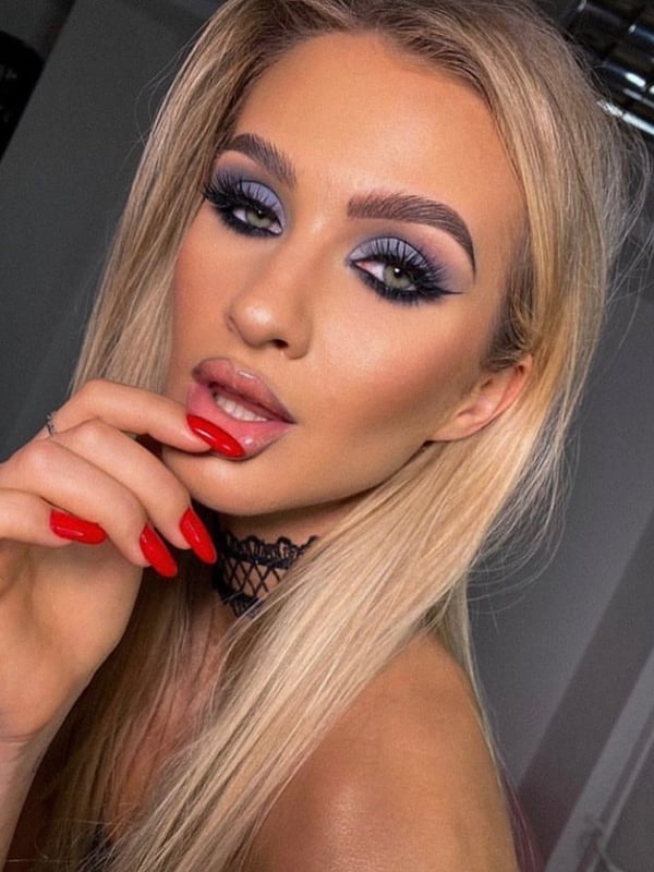 Close-up portrait of a woman with long blonde hair, blue eyeshadow, and red nails wearing a choker necklace.