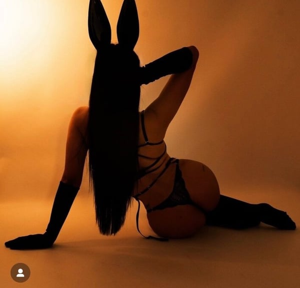 A woman wearing bunny ears and long gloves poses playfully on the floor in a dimly lit room with warm lighting.